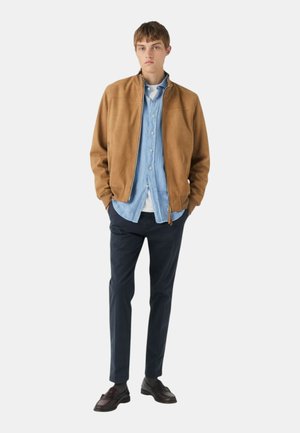 Giacca bomber in suede marrone con zip, camicia di chambray azzurra sotto, pantaloni blu navy e scarpe eleganti scure. Design semplice, vestibilità casual.