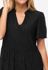 Γυναίκα που φοράει μαύρο φόρεμα με κοντά μανίκια, με διακριτικό μοτίβο διαμαντιού και V-λαιμόκοψη με σχισμή, με το χέρι σηκωμένο κοντά στο πρόσωπό της.