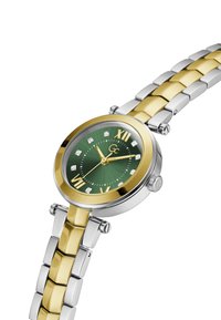 Reloj en tonos dorados y plateados con una esfera redonda verde, marcadores de diamante y números romanos. Pulsera de eslabones metálicos con eslabones rectangulares.