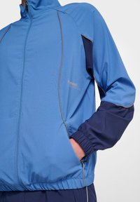 Blå zip-up jacka med marinblå detaljer, texturerat tyg, elastiska ärmslut och sidfickor. Har tunn grå piping längs sömmarna.