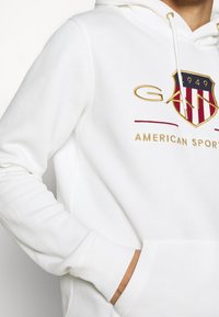 Sudadera blanca de algodón con un bolsillo frontal, que presenta un logo bordado en oro y rojo que representa un escudo y texto.