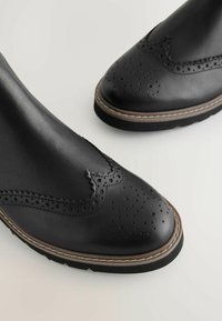 Mustast nahast pahkluu saapad, millel on lõhikuga detailid, brogue-muster, kontrastne õmblus ja tekstuuriga tald.