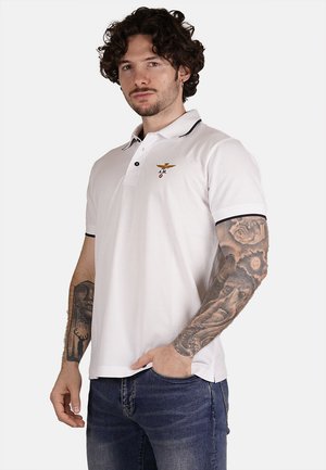 Camisa polo blanca con ribete negro, logotipo bordado en el pecho y corte clásico. Presenta mangas cortas y una textura de algodón suave.