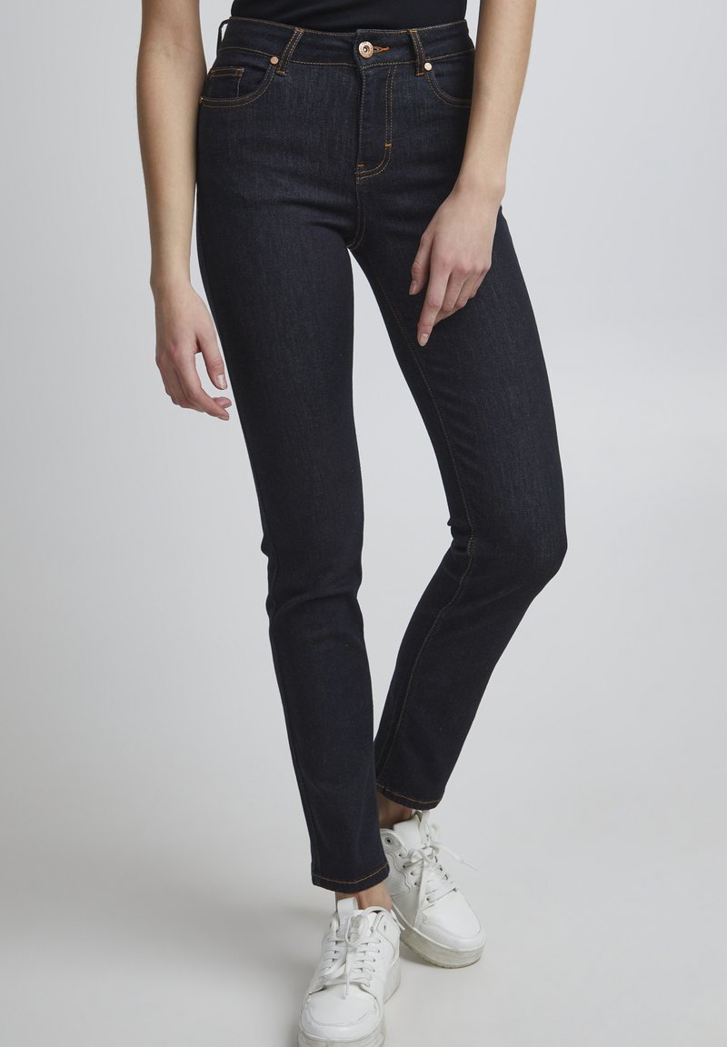 Temno modri skinny jeans iz denima, z oranžnimi šivi, klasičnim pet-pocket dizajnom in visokim pasom. Obuti z belimi športnimi copati.