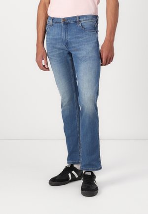 Man wearing light blue straight-leg jeans and black sneakers with white stripes, standing against a plain white background.