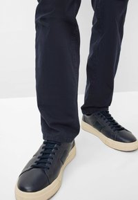 Marine leren sneakers met een gestructureerd oppervlak, een lichtgekleurde rubberen zool en zwarte veters, gecombineerd met donkere broek op een neutrale achtergrond.