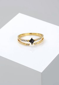 Anello d'oro con una pietra centrale a forma di diamante nero, affiancato da piccoli cristalli trasparenti, con un design a fascia divisa e slanciata.