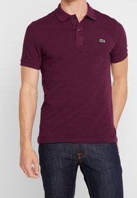 Burgundské polo tričko vyrobené z bavlny, s tříknoflíkovou légou, krátkými rukávy a zeleným logem na hrudi. Hladká textura.