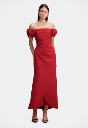 Femme portant une robe rouge longue jusqu'au sol, épaules dénudées, manches bouffantes et talons ouverts noirs, debout devant un fond uni.
