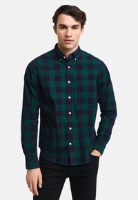 Groen en marineblauw geruit shirt met button-down kraag en een borstzak, gemaakt van een gestructureerde stof. Model draagt het gestopt in zwarte broeken.