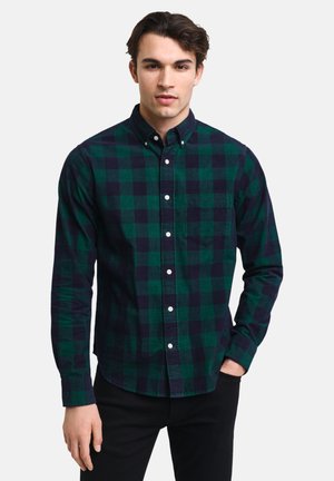 Groen en marineblauw geruit shirt met button-down kraag en een borstzak, gemaakt van een gestructureerde stof. Model draagt het gestopt in zwarte broeken.