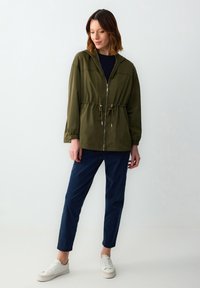 Giacca a zip verde oliva con cappuccio, vita cintrata, polsini elasticizzati e tasche inclinate, abbinata a pantaloni blu navy e sneakers bianche.