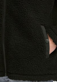 Black fleece jacket with a textured surface, zip closure, and side pockets. Features a black button accent on the pocket opening.
