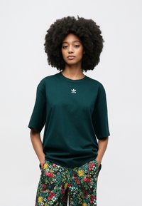 Mørkegrøn bomulds-T-shirt med korte ærmer, med en lille hvid adidas-logo. Sat sammen med mønstrede blomstrede bukser i levende farver.