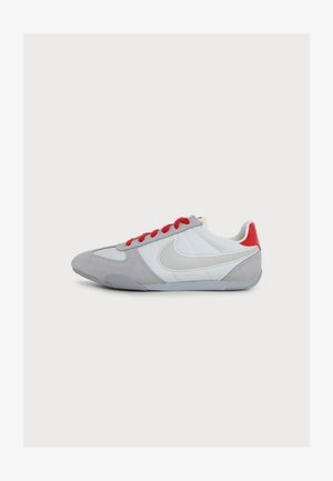 Nízkoprofilová teniska so svetlosivými a bielymi panelmi, červeným detailom na päte, červenými šnúrkami a veľkým bielym logom Nike swoosh na boku.