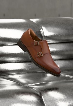 Chaussure à double boucle en cuir marron flottant au-dessus d'un canapé capitonné argenté métallique brillant.