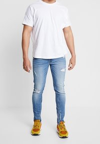 Camiseta de algodón blanca, jeans ajustados rasgados de color azul claro y zapatillas amarillas con acentos naranjas y negros. Presentación de un atuendo casual.