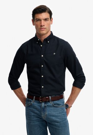 Mann mit dunklem Haar, der ein schwarzes Hemd mit Knöpfen und hochgekrempelten Ärmeln, blaue Jeans, braunen Gürtel und Armbanduhr trägt, steht mit verschränkten Händen hinter dem Rücken.