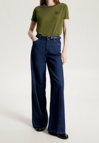 Olivgrön bomulls-t-shirt med uppvuxna ärmar och broderad logotyp, matchad med mörkblå högmidjade vida jeansbyxor och ett svart bälte.