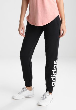 Osoba nosící černé tepláky Adidas s bílým logem na dolní části nohy, růžové tričko a bílé sportovní boty, stojící na světlém pozadí.