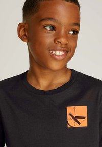 T-shirt nera in cotone con un logo quadrato arancione con le lettere "CK" in nero, collo rotondo e maniche corte.