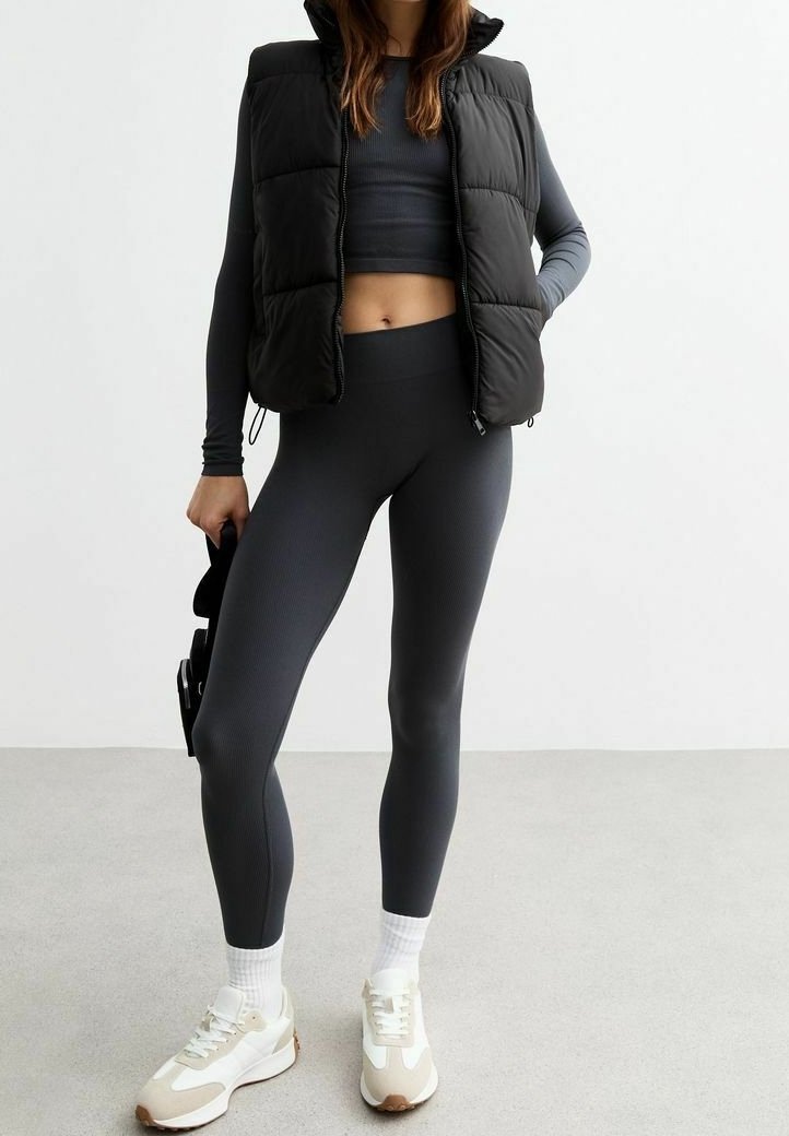 Černá nadýchaná vesta přes tmavě šedé crop top, doplněná o šedé žebrované legíny a bílé tenisky s hnědými akcenty.