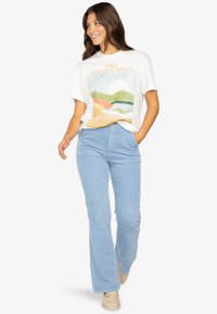 Hvid grafisk t-shirt med farverigt bjergdesign, parret med lyseblå kordfløjspantser og beige sneakers. Afslappet pasform.