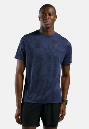 Fit man wearing dark blue patterned short-sleeve athletic shirt and black shorts, standing against a plain white background.