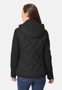 Chaqueta de plumas negra con textura acolchada, capucha y mangas ajustadas estilo suéter, combinada con jeans azul oscuro. Se muestra vista trasera.