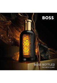 Schwarze Glasparfümflasche mit goldener Kappe, auf der "BOSS" in kräftigen Lettern geprägt ist. Vor einem dunklen, strukturierten Hintergrund mit natürlichen Akzenten.