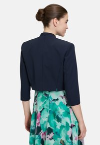 Skórzana granatowa kurtka o 3/4 rękawach, o prostym kroju i gładkiej fakturze. Nosi ją osoba na sukience w kwiatowy wzór w odcieniach zieleni i różu.