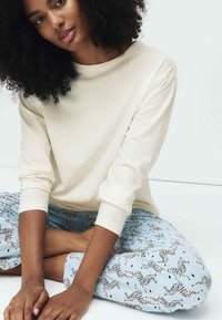 Lysebeige langærmet skjorte kombineret med blå pyjamasbukser med sort-hvidt zebra mønster; siddende positur med arme krydset.