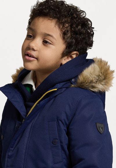 Σκούρο μπλε παρκά με κουκούλα επενδεδυμένη με γούνα, κίτρινο φερμουάρ και κουμπιά. Διαθέτει λογότυπο στη μανσέτα.