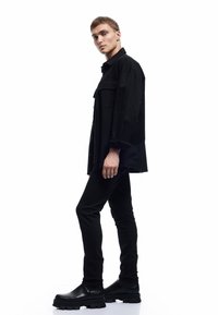 Camisa negra oversized con bolsillos frontales, combinada con pantalones negros ajustados y botas negras de suela gruesa. Tejido suave y corte recto.