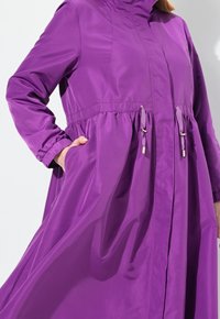 Manteau léger violet avec un col haut, une taille à cordon de serrage et des poignets élastiques. Comprend des poches latérales et un design fluide et aérien.