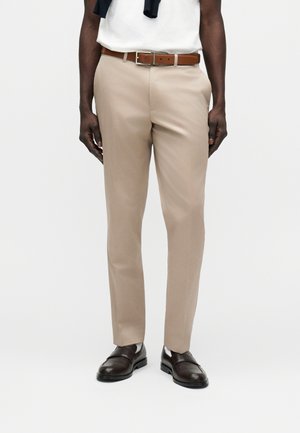 Άνδρας που φοράει μπεζ ραμμένο παντελόνι με καφέ ζώνη και σκούρα καφέ δερμάτινα παπούτσια, στέκεται μπροστά σε ένα απλό φόντο.