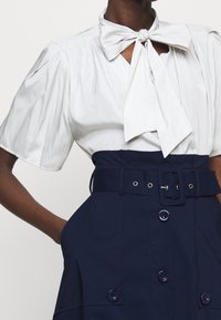 Blusa bianca con un fiocco al collo, maniche corte a palloncino e una gonna a vita alta blu navy con una cintura larga e bottoni.
