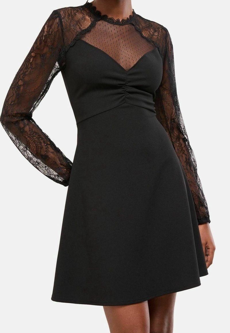 Robe noire avec manches longues en dentelle transparente, haut en résille avec pois, buste froncé et jupe évasée portée par une personne debout.