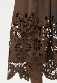 Καφέ ύφασμα με κεντήματα με λουλουδάτα κοψίματα και λεπτομέρειες από δαντέλα κατά μήκος του τελειώματος ενός ενδύματος.