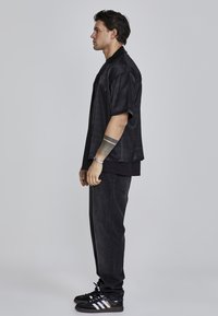 Camisa negra de manga corta con un patrón texturizado, combinada con jeans oscuros y zapatos negros con rayas blancas y moradas.