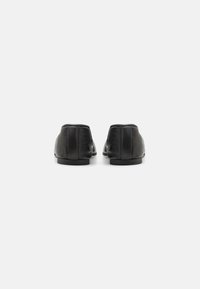 Chaussures en cuir noir avec une texture lisse, un profil bas et un design minimaliste. L'arrière présente des détails de couture subtils et une semelle plate.