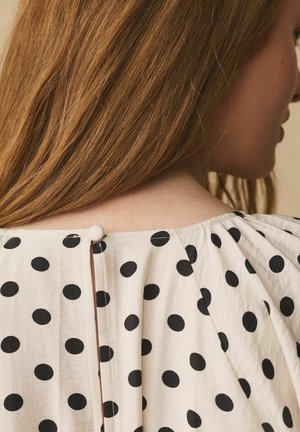 Woman with long brown hair wearing a white blouse with large black polka dots, buttoned at the back near the neck.