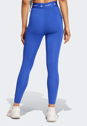 Personne debout portant des leggings taille haute bleu vif avec une ceinture "TECHFIT" et des chaussures de sport blanches, vue de dos.