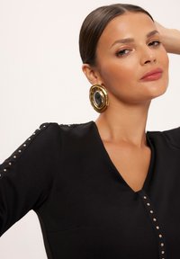 Czarna bluzka z dekoltem w serek z złotymi ćwiekami na ramionach i centralnym detalem w postaci guzika, w zestawieniu z dużymi, złotymi i czarnymi kolczykami.