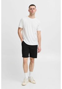T-shirt en coton blanc à manches courtes, associé à un short noir. Le mannequin porte des chaussettes blanches et des baskets crème, se tenant devant un fond uni.