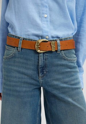 Brauner Ledergürtel mit gehämmerter goldener Schnalle, getragen über blauen Jeans und in ein hellblaues Hemd mit Knöpfen gesteckt.