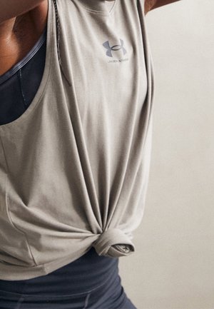 Osoba w jasnoszarym bezrękawniku Under Armour związanym w talii, na ciemnym sportowym biustonoszu i legginsach, podnosi ręce ponad głowę.
