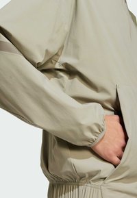 Ľahká béžová bunda s elastickými manžetami, voľným strihom, prednou kapsou a hladkým, textúrovaným materiálom pre pohodlie a ľahký pohyb.