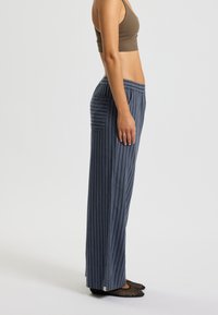 Pantaloni da pigiama a righe blu con vestibilità ampia, vita elastica, tasca laterale e design a gamba dritta. Il tessuto appare morbido e leggero.