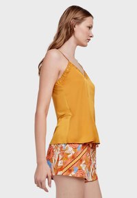Top sin mangas amarillo con escote en V y borde decorativo de ojetes, combinado con pantalones cortos estampados que presentan diseños florales y geométricos en tonos naranja y azul.